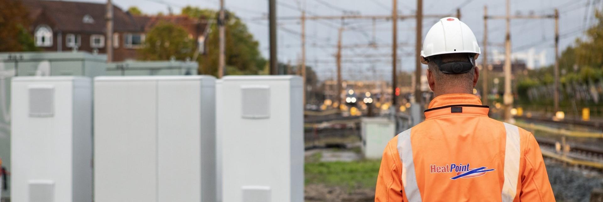 Een Heatpoint medewerker in een oranje veiligheidsvest en witte helm, met “HeatPoint” erop, staat voor een spoorweg. Er staan meerdere wisselverwarmingskasten aan de zijkant.